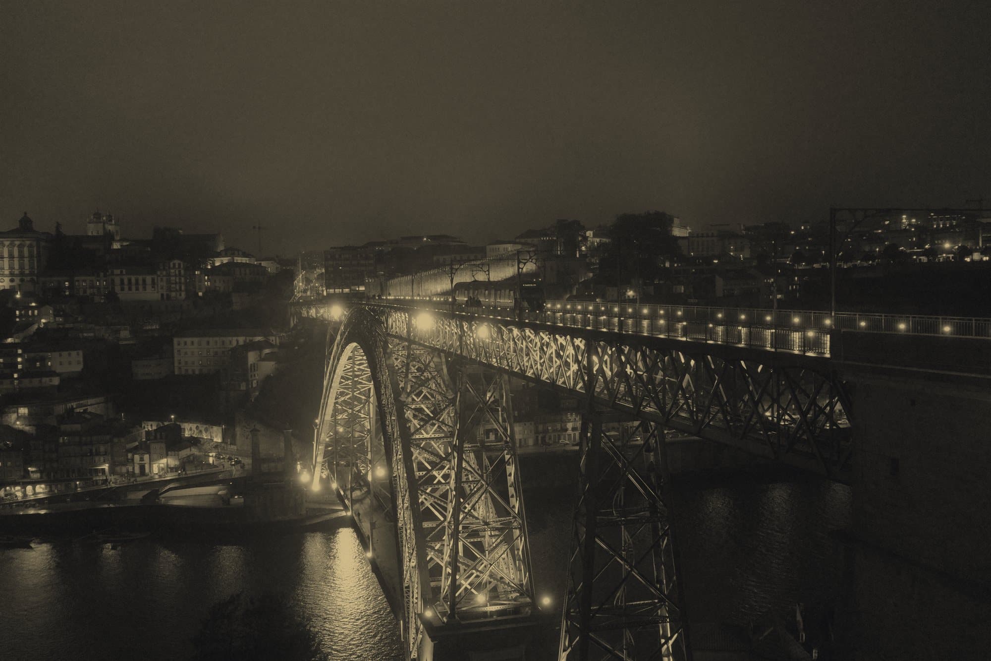 An illuminated steel truss bridge spanning a river at night, its triangulated framework lit against the dark sky.